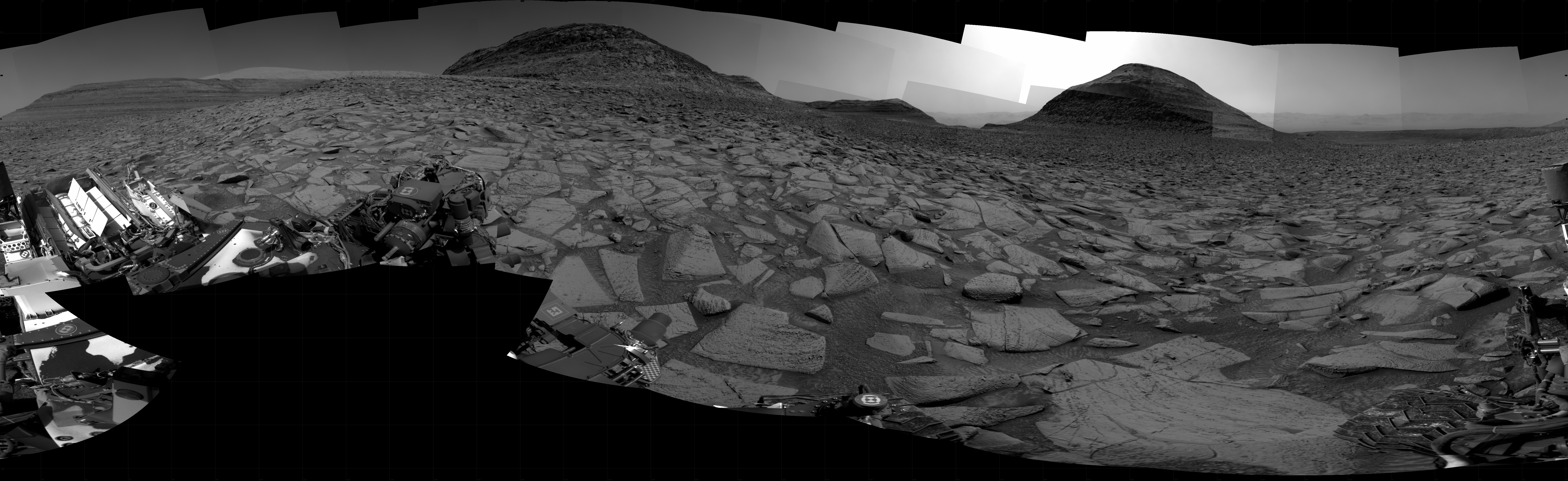 A grayscale panorama of the Martian surface, possibly at night or in twilight, shows a wide field of flat, dark gray terrain dotted with flat, angular, medium-sized rocks, lighter gray, stretching into the distance where a pair of hills rise from the ground on the horizon. A bright light illuminates a small part of the sky, off the frame and above the smaller hill on the right. A third hill begins to slope upward on the horizon, at the right edge of the frame. Portions of the Curiosity rover are visible at the bottom of the image, including a wheel visible in the bottom center of the frame, and two others in the lower right corner.