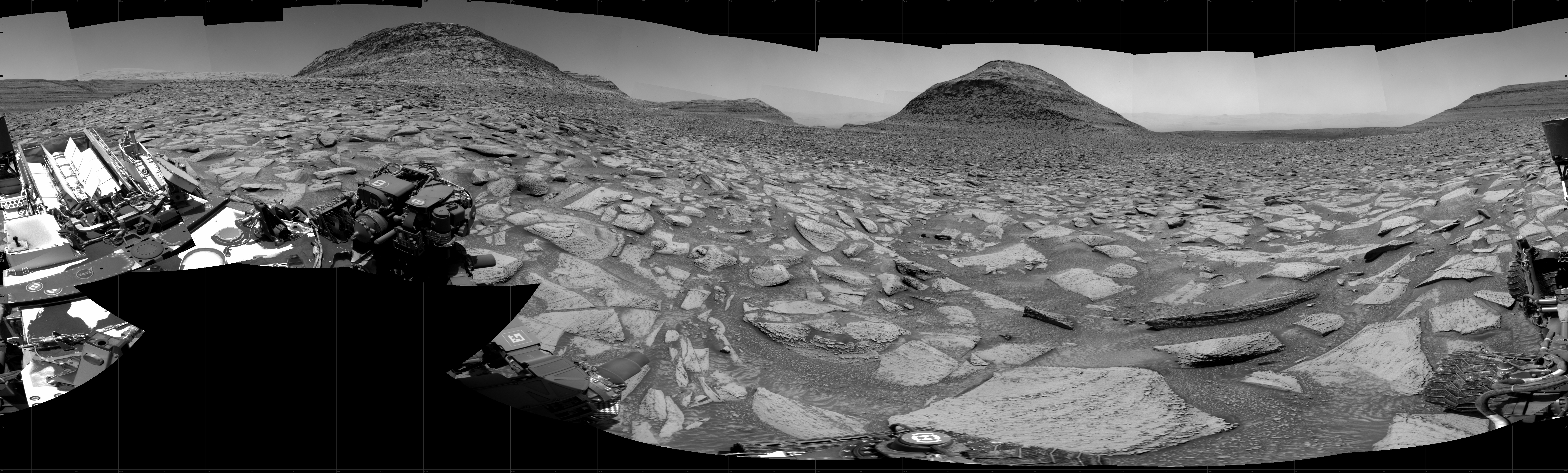 A grayscale panorama of the Martian surface shows a wide field of flat, dark gray terrain dotted with flat, angular, medium-sized rocks, all in lighter gray, stretching into the distance where a series of four hills rise from the ground, left to right on the horizon. Portions of the Curiosity rover are visible at the bottom of the image, including two wheels visible in the lower right corner and the top deck of the rover on the left side of the image, including a NASA logo. The terrain at image left slopes downward off the frame, and more distant areas are visible beyond.