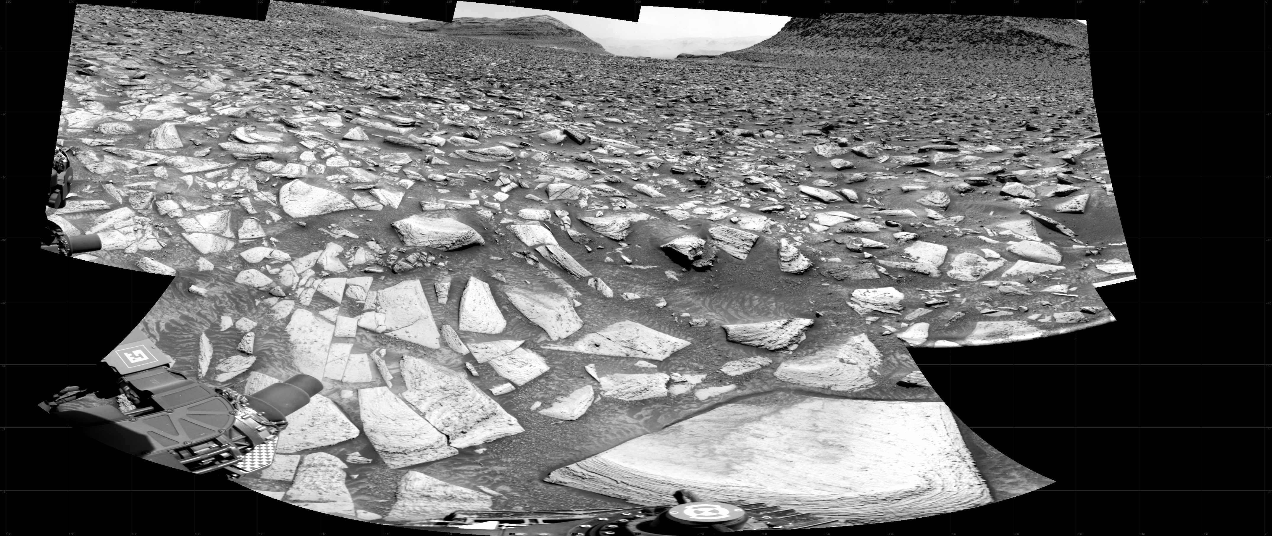 A grayscale photograph of the Martian surface shows very uneven rocky terrain, with bright-toned, flat, lined and multi-angled rocks covering the surface, with darker soil in between, looking like the entire foreground stretching off into the distance had shattered. On the horizon, a pair of rocky hills rise at the center of the frame. Portions of the rover are visible at the bottom and left side of the frame.