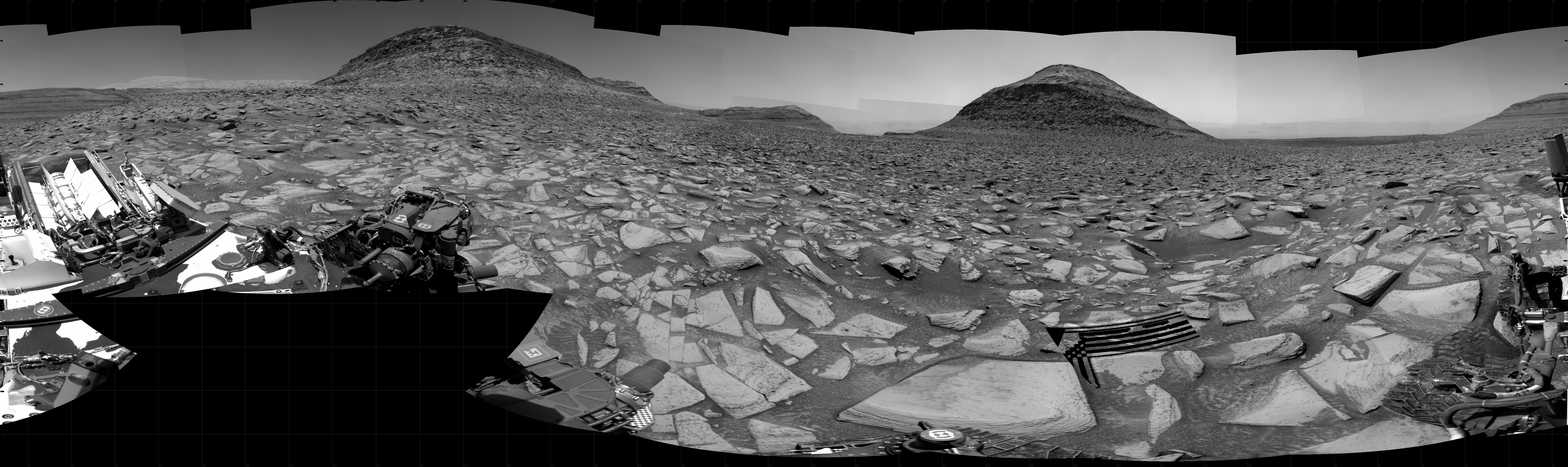 A brightly lit, grayscale panorama of the Martian surface shows very uneven rocky terrain, with bright-toned, flat, lined and multi-angled rocks covering the surface, with darker soil in between, looking like the entire foreground stretching off into the distance had shattered. On the horizon, four hills rise from the ground — a small one at the center of the image, two larger ones on either side of it, and a fourth on the right side of the image that angles upward off the frame. Portions of the rover are visible at the bottom of the frame, from one corner of the other, including one wheel at bottom center, and two others in the lower right corner, along with visible wheel tracks on the ground there.