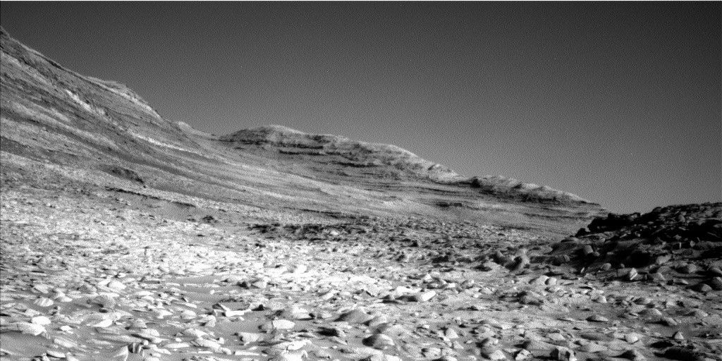 A grayscale photo of a Martian landscape shows very rough and rocky terrain extending from the foreground toward large features in the distance; they are rocky, looking like they are composed of multiple stacked layers rising from the ground, sloping gently down toward the right side of the image. On the far right, a smaller outcrop, shadowed and darker than the rest of the scene, its surface encrusted with small, jagged rocks.