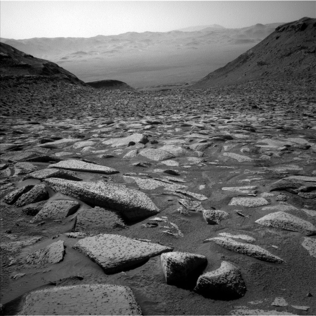A grayscale image from the Martian surface shows very rocky, medium gray terrain in the foreground leading to V-shaped gap in the distance between two gently sloping hills on either side of the image. The foreground is covered in medium-sized rocks of many shapes and angles pushing up from the soil. Beyond that the ground recedes and dips far into the distance toward a crater floor, with the crater walls hazy but visible very far on the other side.