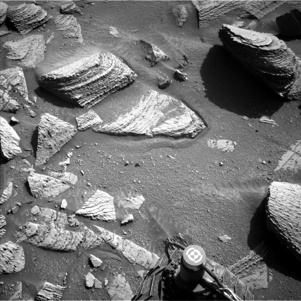A grayscale photograph of the Martian surface from the Curiosity rover captures medium-gray, coarse soil in front of the rover, with rocks everywhere protruding up through it. They are lighter-toned than the ground, some rounded, others looking like cracked, smooth plates. The largest rocks look like they’re built up from stacked layers. A part of the rover is visible in the frame, at bottom center, including one of its wheels.