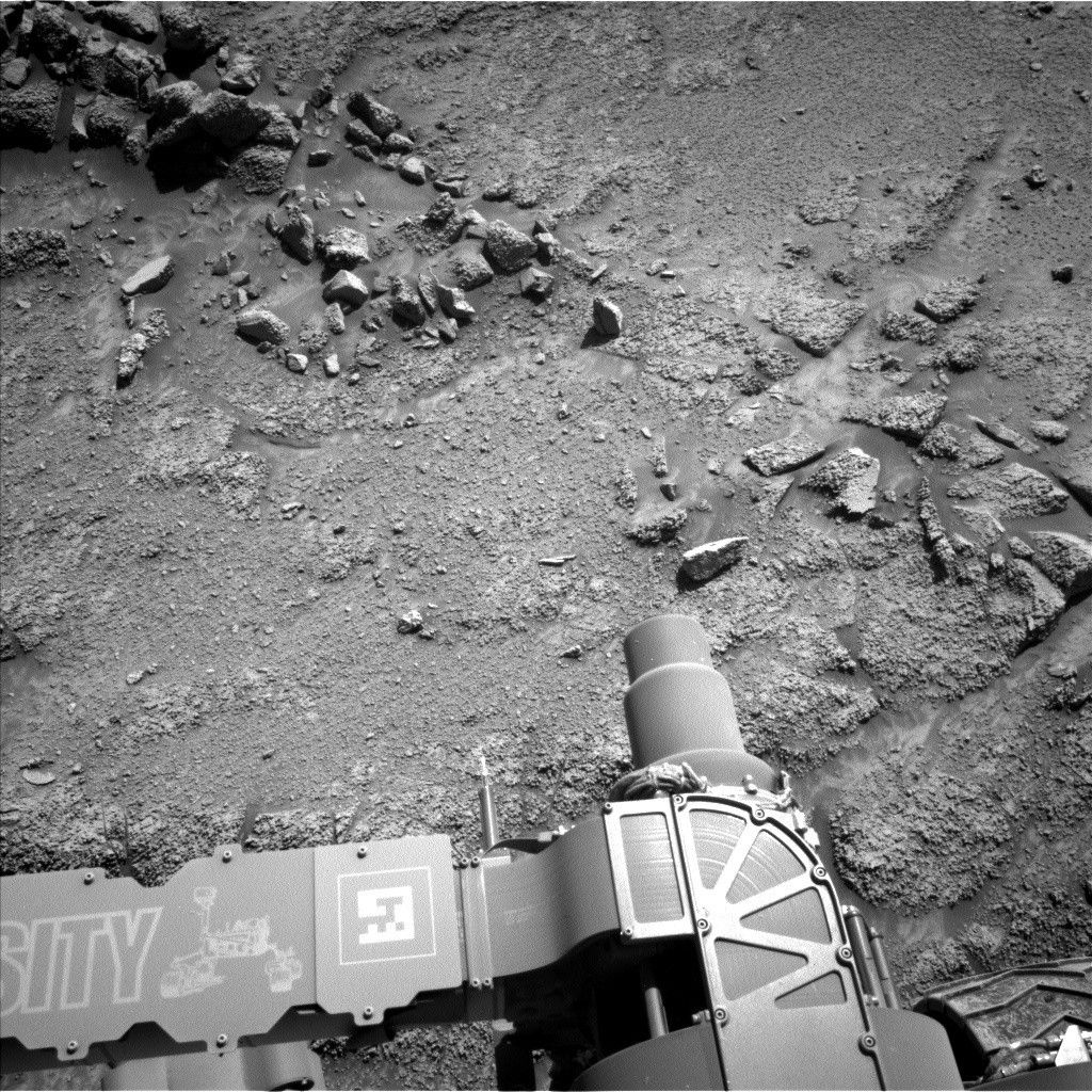 A grayscale photograph of Martian terrain in front of the Curiosity rover shows uneven, medium-gray ground with numerous rocks of many sizes scattered around, mostly running in a diagonal line from the upper left corner of the image to the center-right edge of the frame. The bottom of the frame shows parts of the Curiosity rover, with a wheel visible in the lower right corner of the frame, and part of its robotic arm running from the lower-left corner to the bottom center of the image, with a nameplate imprinted with “Curiosity” outlined in white, all-capital letters, and to the right of that a line drawing of the rover. A small crescent-shaped rock is visible on the ground above the “SI” printed on the arm.