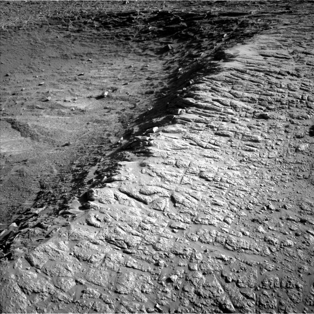 A grayscale photo from the Martian surface shows rough terrain covered in small rocks, divided across the middle of the image by a ridge running from the lower left corner of the frame to the upper right corner. The ground to the right of the ridge is relatively flat, while to the left it falls away from the ridge into a depression. The entire scene is light to medium gray, except for a very dark shadow along the left side of the ridge.