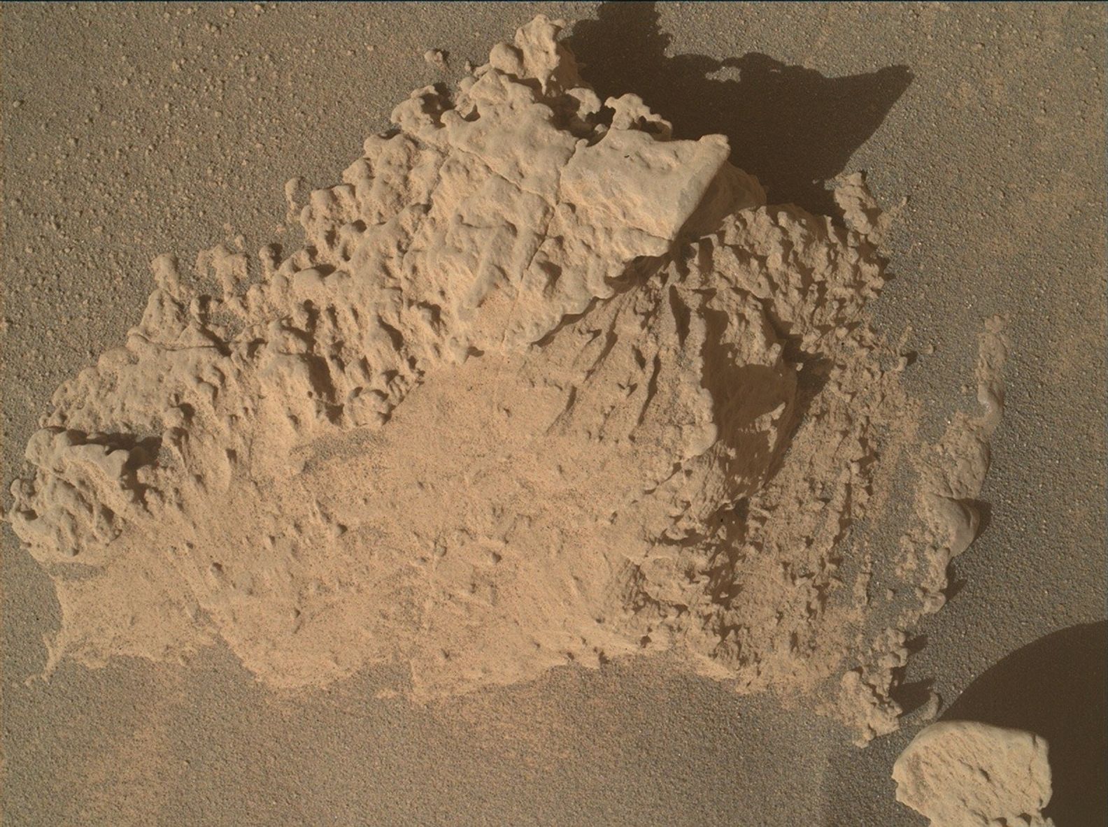 A color image from the Martian surface shows a close-up of a pale orange-tan rock sticking out above the sandy soil around it, dominating the center of the image. The right side of the rock appears lower, with rough-edged layers resembling a wide staircase rising from ground level toward the center of the rock. The upper left side of the rock appears to be higher, with pockmarks and missing areas that look smoother than the rest of the rock.