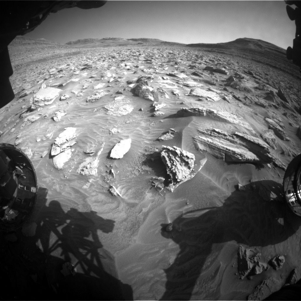 A grayscale, wide-angle photo from the Martian surface shows very rocky terrain in front of the rover leading to the horizon, where a series of distant buttes and gently sloped hills rise from the ground. Rocks of various shapes and sizes jut out from the soil in the foreground, some flat and smooth while others are heavily textured, and several show distinct layers. The soil surrounding them appears finely textured, and in many places looks wavy, as if sculpted at some point by wind or water.
