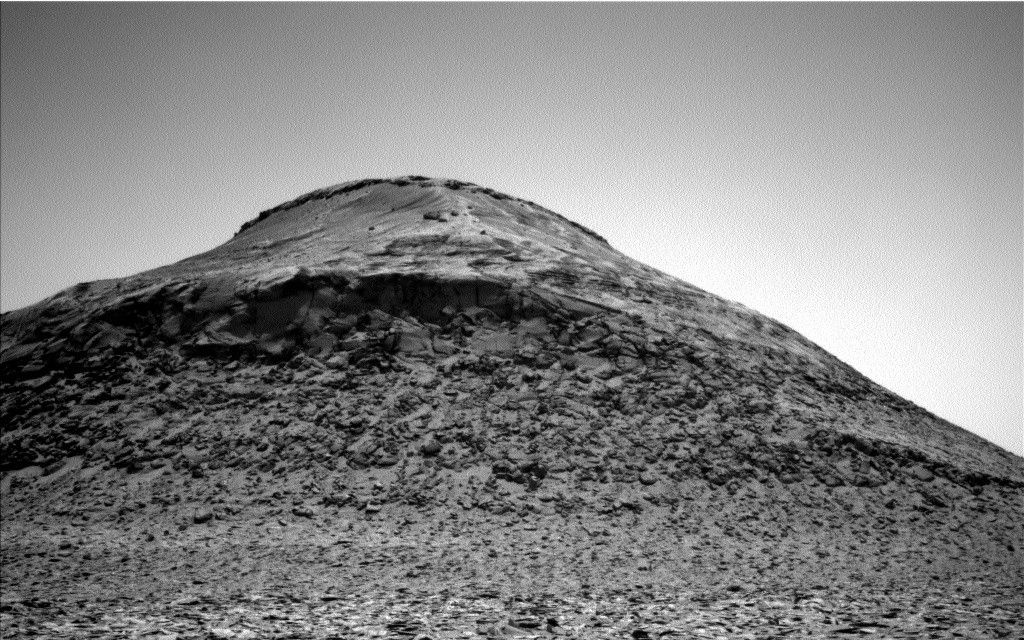 A grayscale image from the Martian surface shows a gently sloping hill in the background at the upper center of the photo, with fairly smooth, medium gray terrain around its peak. In the foreground leading up to the hill, the terrain is much rougher, covered in a multitude of small rocks. The sky above is clear and a very pale gray.