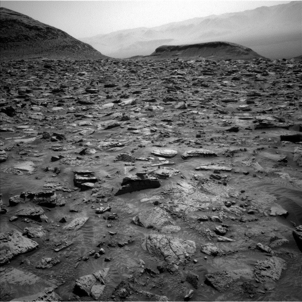 A grayscale image from the Martian surface shows very rocky, medium gray terrain in the foreground leading to a gently sloping hill on the horizon at left, and a smaller butte at image center. The ground is covered in medium-sized rocks of many shapes and angles pushing up from the soil. Prominent in the center foreground is a medium-sized, wedge-shaped rock that points toward the right and is much darker than the surrounding terrain.