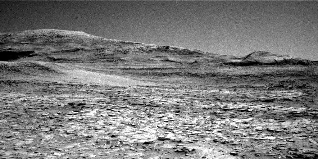 A grayscale photo from the Martian surface shows rocky ground from the front of the image to a horizon line where a series of gently sloped hills rise up.