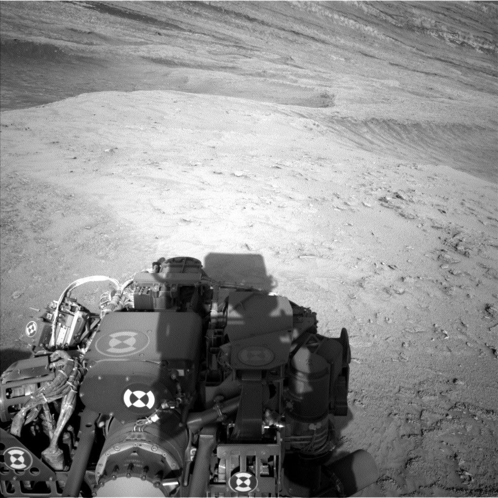 A grayscale photo shows Martian terrain, which appears medium gray throughout. In front of the Curiosity rover, the ground from the bottom to the middle of the frame appears relatively smooth, with a few scattered small rocks poking above the surface. In the upper part of the image, the terrain appears to slope downward from left to right, with ridges running down along that same direction. Part of the rover is visible in the lower-right corner of the image, casting a shadow on the ground.