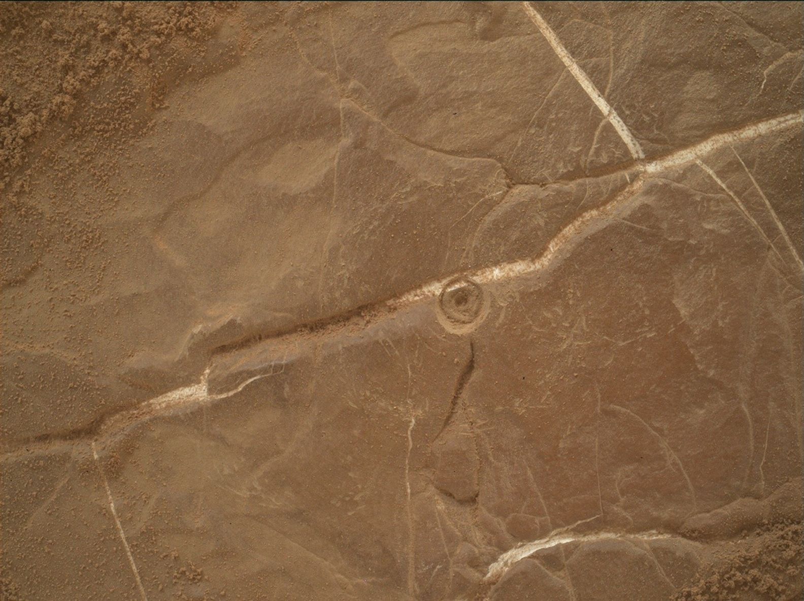 A color image from the Martian surface shows a close-up of brownish-orange ground, flat but uneven, with whitish grooves crisscrossing the surface. At center a small, bullseye-shaped indentation has been dug into the rock.