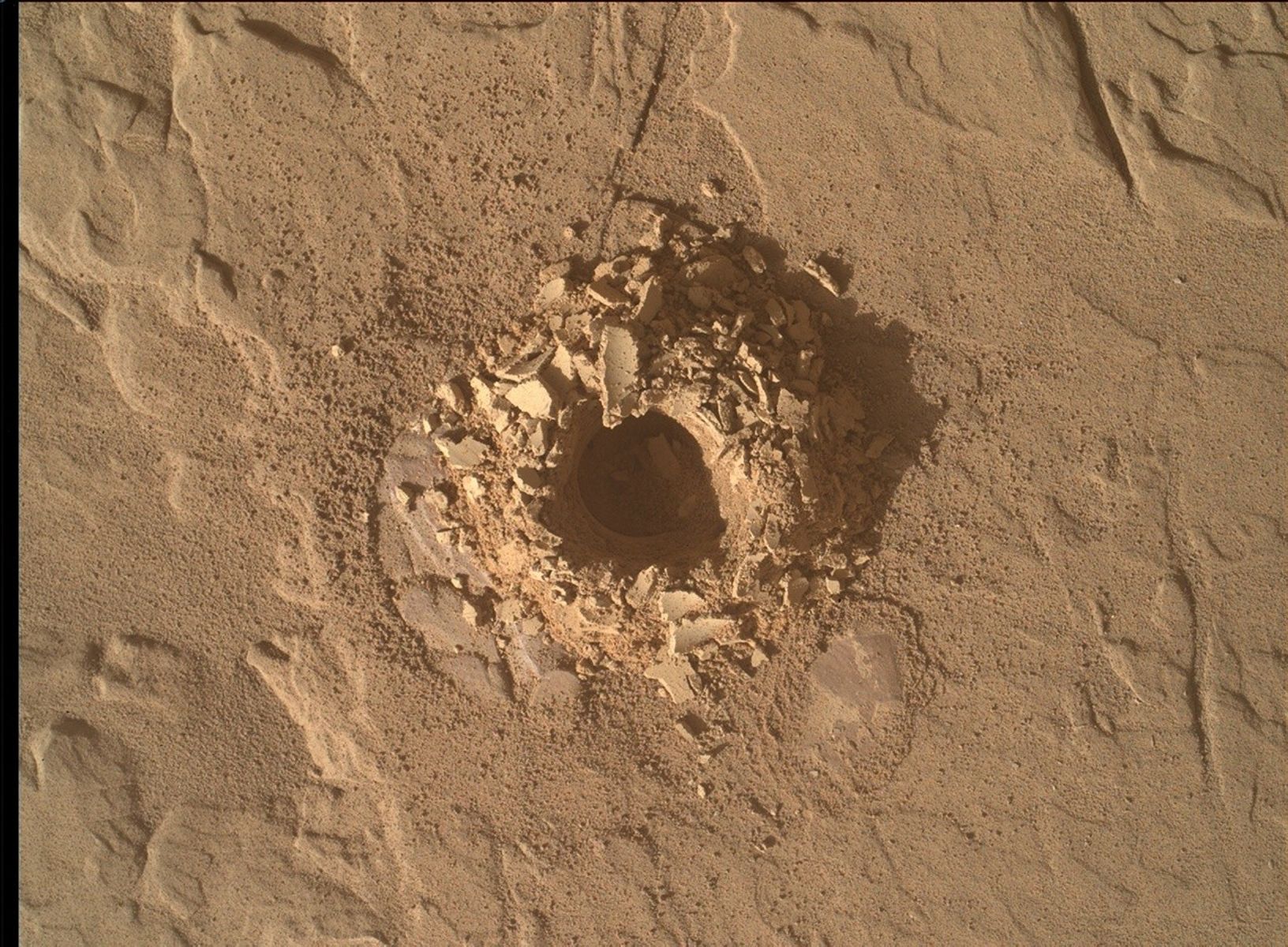 A color close-up photo of the Martian surface shows flat, mostly smooth, tan-orange ground, marked with a few shallow grooves and indentations. At the center is a hole cored into the ground, surrounded by material dug out from the hole, which looks like a mixture of soil and sharp flakes or shards of rock. Those are similarly colored to the surrounding ground.