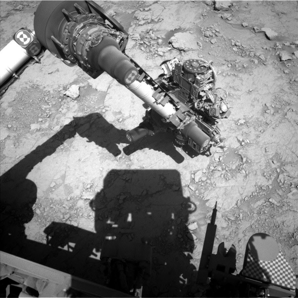 A grayscale photo from the Martian surface shows the Curiosity rover investigating terrain that is mostly flat, with small- to medium-sized rocks scattered throughout the area, all of it light- to medium-gray. The rover’s robotic arm dominates the center of the image, as it is reaching down toward the surface. Other parts of the rover are visible at the bottom of the frame, parts in sunlight and other parts shadowed, and the shadow of the rover’s large, block-shaped Mast Head is also visible.