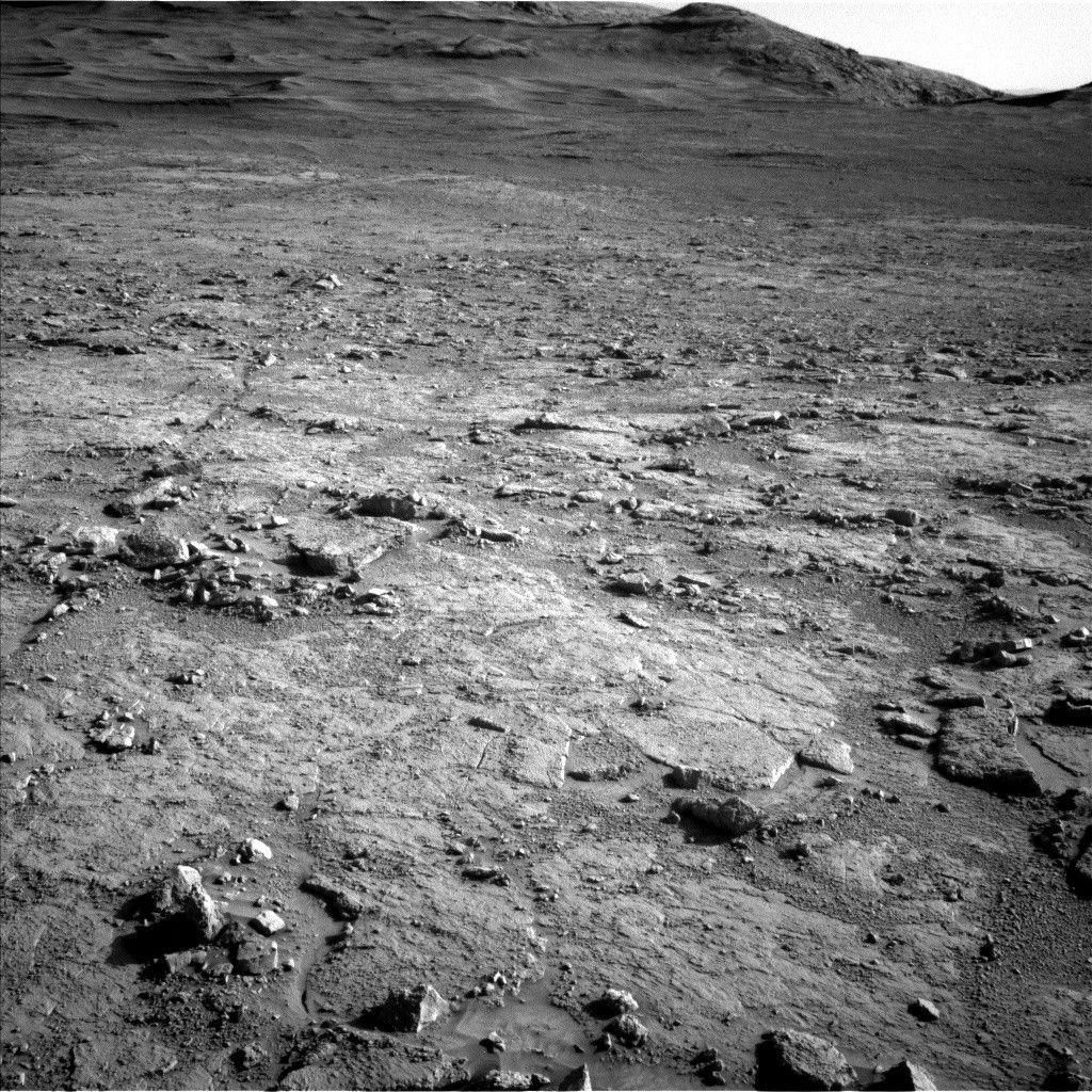 A grayscale photograph of the Martian surface from the Curiosity rover shows a mostly flat, stony field, with rough and jagged medium-sized rocks scattered around, extending to hills in the background, all medium to dark gray. The hills appear smoother overall, with waves on the left side that look like windblown dunes.