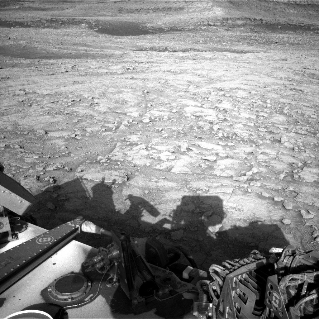 A grayscale photograph from the Martian surface shows rough terrain covered in a multitude of small- to medium-sized rocks, resting atop timeworn surface features, all colored a fairly light gray. Part of the Curiosity rover, and a shadow it is casting, are visible at the bottom of the frame.