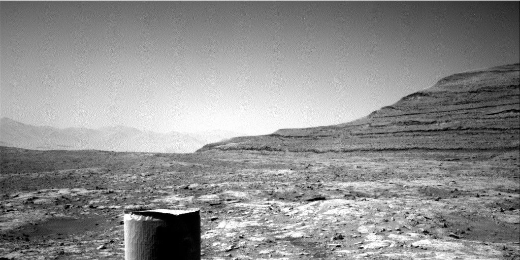 A grayscale photo of a Martian landscape shows very rough, rocky, and cracked terrain extending from the foreground to a horizon line in the distance, across the middle of the frame. From the midpoint of that horizon line, a layered hill rises toward the upper right side of the image. In the far distance, a hazy stretch of dunes and hills is visible. And a cylindrical portion of the rover is visible in the foreground, extending up from the bottom edge of the frame.