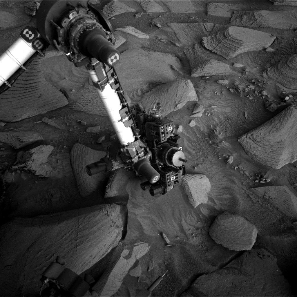 A grayscale photo from the Martian surface shows the Curiosity rover’s robotic arm reaching down to investigate a targeted rock. The surrounding terrain is very rocky and uneven, with stones of varied shapes and sizes pushing up out of the ground, everything a similar dark shade of gray. The soil between the rocks appears wavy in many places, as if sculpted by wind or water, and most of the rocks look like they have formed from horizontal layers stacking on one another, in the relatively short elevation they rise from the surface, looking like 3D versions of hills indicated on a topographic map. The rover’s arm extends down to the center of the image from the upper left corner of the frame where, at a pivot point, it bends at roughly 90-degree angle toward the left side of the image and out of the frame. The arm itself is bright-toned, nearly white, with a metallic sheen; the pivot point and the boxy suite of instruments at the end of the arm near the ground are mainly dark gray or black, with multiple knobby protrusions.