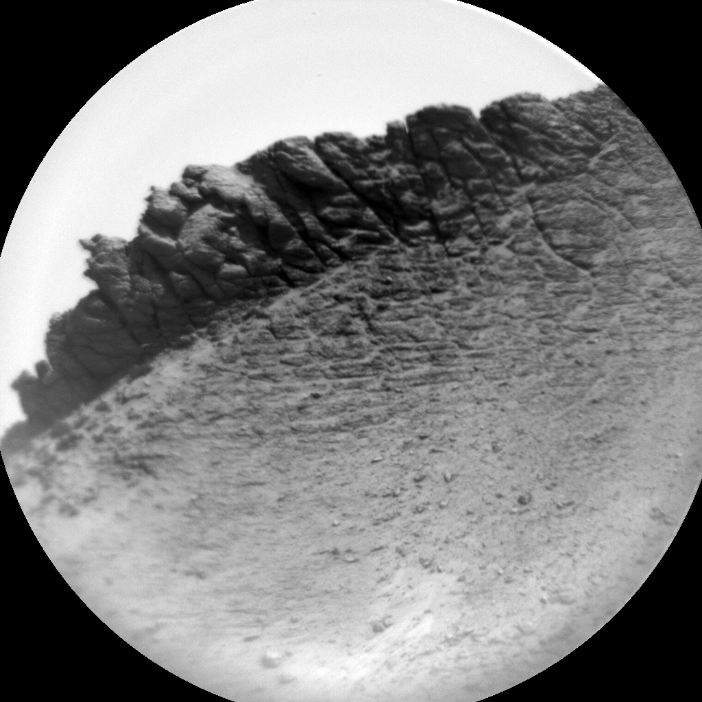 A circular grayscale image from the Martian surface is skewed slightly left, so the horizon line runs from about the 8 o’clock position on a clock face to about 2 o’clock. Gritty, gravelly terrain in the foreground leads to a spiky, ragged outcrop with vertical ridges and grooves rising on the horizon.