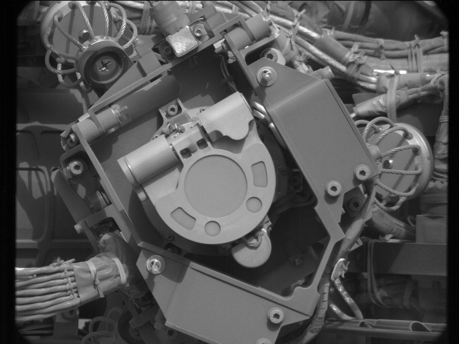 A grayscale close-up image of an instrument on the Mars rover Curiosity, showing at the center of the image a U-shaped flat piece of metal face the camera, with its rounded bottom pointing toward the lower right corner of the image. It’s surrounded by metal plates at different angles, some parallel to the plane of the image, others perpendicular, and most of them secured with hex nuts. Braided bunches of cable are twisted behind these other sections, leading into the instrument.