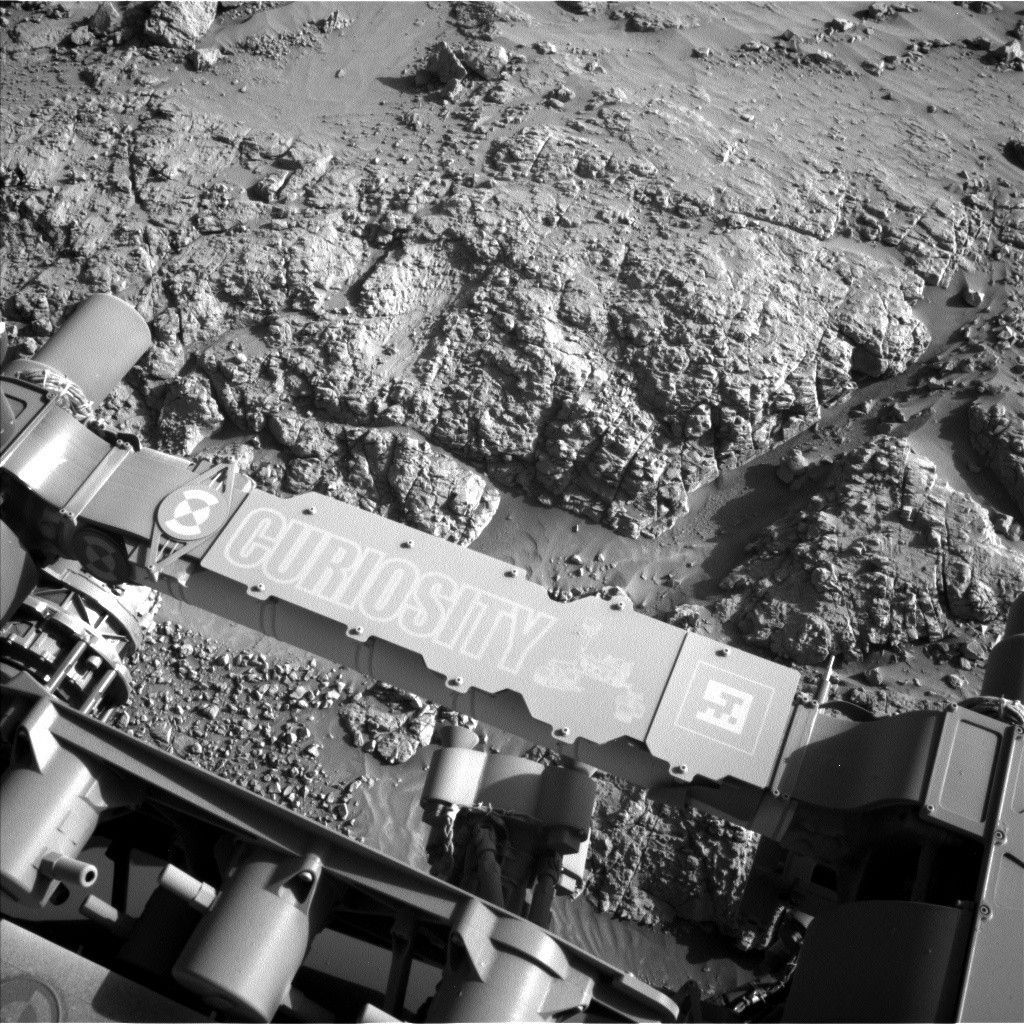 A grayscale photograph of Martian terrain in front of the Curiosity rover shows very bumpy, uneven ground with large, polygonal sections of rock on the surface separated by gouges in the soil. The bumpy area and a smoother patch just beyond that all appear shiny. The bottom of the frame shows parts of the rover, running from the middle left edge to the lower right corner of the image, including part of its robotic arm which carries a nameplate imprinted with “Curiosity” outlined in white, all-capital letters, and to the right of that a line drawing of the rover.