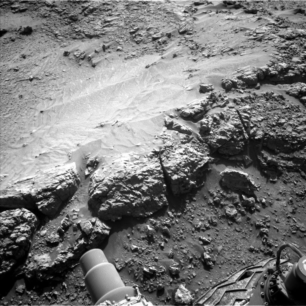 A grayscale photograph of Martian terrain in front of the Curiosity rover shows a ledge of flat, rough ground that appears to drop off just in front of the rover. Portions of the rover are visible at the bottom of the frame, including a wheel in the lower-right corner. An area of the ground ahead is smoother than the rest, looking like small, wavy sand dunes. Many of the small to medium sized rocks surrounding this patch have trails of sandy soil running off to their right, as if wind or water had flowed from left to right and sculpted these pointed tails of runoff.