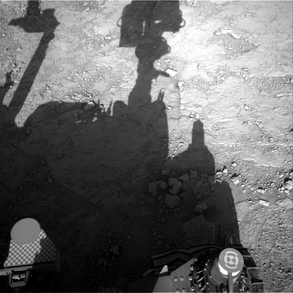 A grayscale photo looking down at the Martian surface, showing very rough terrain in front of the Curiosity rover. Light gray, gravel-covered rocks are in bright sunlight in about half the frame, extending up diagonally toward the upper right corner of the image. A shadow silhouette of the rover covers most of the rest of the image, with its mast head and robotic arm appearing very prominently. Portions of the rover itself are shadowed but visible at the bottom of the image.