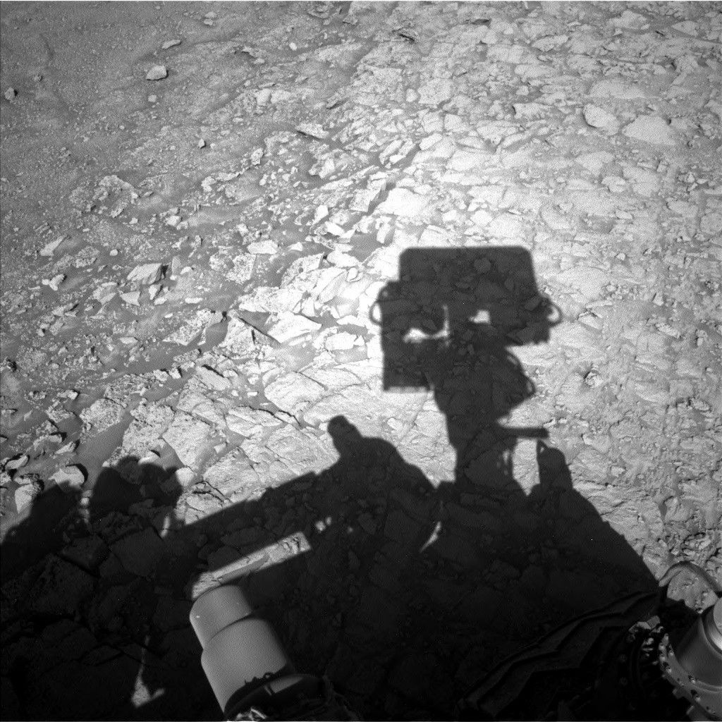 A grayscale photo from the Martian surface shows a rough, rocky landscape in medium gray, with a shadow of the Curiosity rover’s boxy masthead showing at the center of the frame, and the shadow of its body covering some of the bottom of the frame. Along an uneven vertical border from the top to the bottom of the image, the texture of the terrain is separated from more ground-up soil and rock of many sizes, while the right side is covered in larger, flatter rocks, with numerous cracks and crevices separating them from each other.