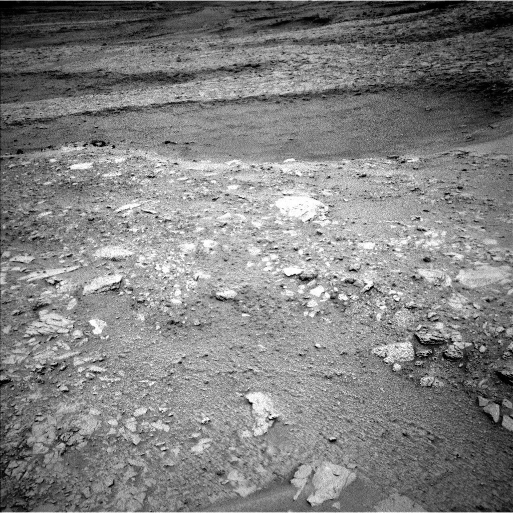 A grayscale image from the Martian surface shows dark gray, very rocky terrain. In the foreground, whitish rocks are surrounded by gray soil, while in the middle of the frame a smoother area looks like sand, and beyond that the terrain turns rocky again, sloping downward from the upper right corner of the frame to the upper left side of the image.