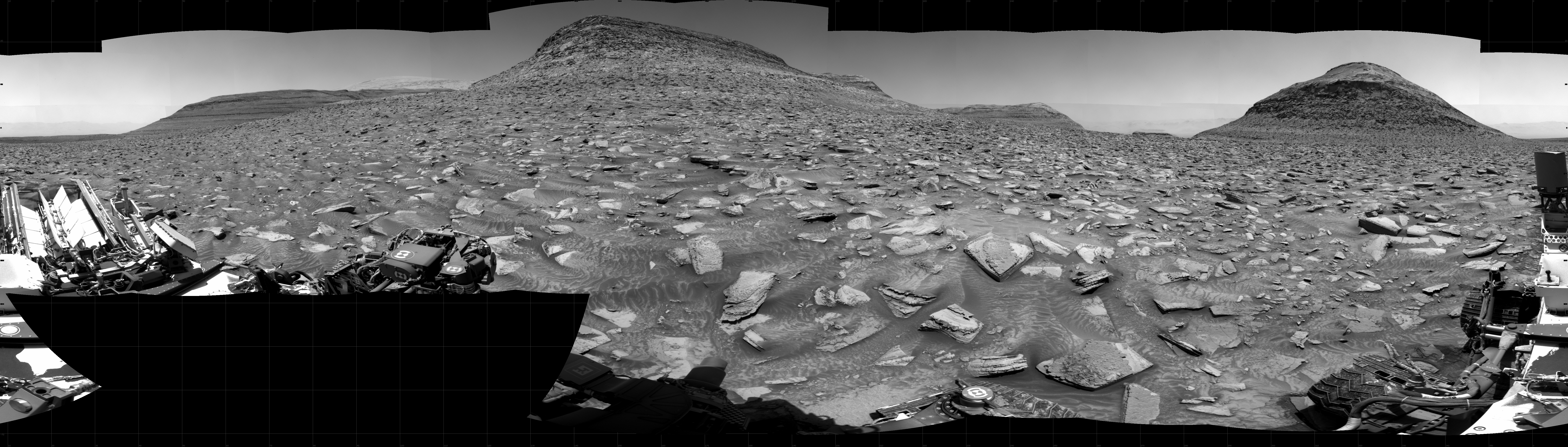 A brightly lit, grayscale panorama of the Martian surface shows very uneven rocky terrain, with bright-toned, flat, lined and multi-angled rocks covering the surface, with darker soil in between, looking like the entire foreground stretching off into the distance had shattered. On the horizon, a hill rises at the center of the image, its peak softened like a timeworn pyramid, with two flat mesas extending to either side at the base. Portions of the Curiosity rover are visible at the bottom of the frame, from one corner to the other, including one wheel at bottom center, and two others in the lower right corner, along with undulations throughout the soil nearby, looking in some places like wheel tracks and in others like sand scalloped by wind or water.