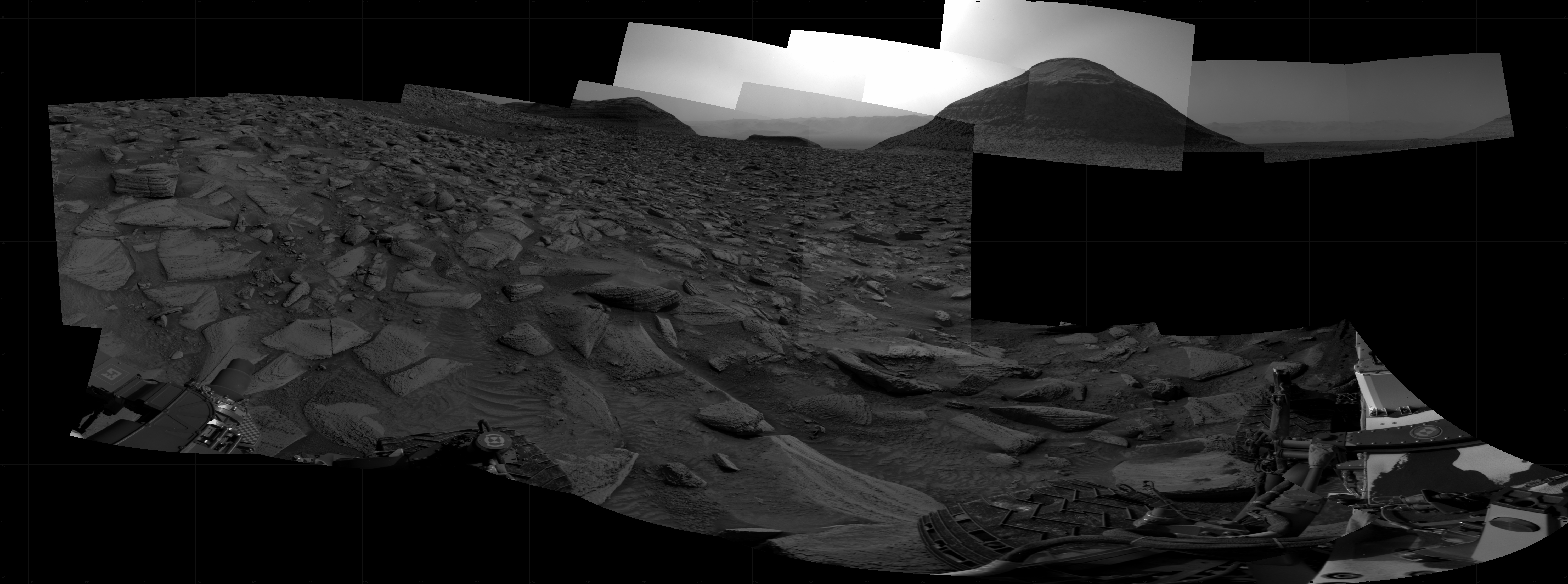 A grayscale panorama of the Martian surface, possibly at night or in twilight, shows a wide field of flat, dark gray terrain dotted with flat, angular, medium-sized rocks, lighter gray, stretching into the distance where a pyramid-shaped hill rises from the ground on the horizon, just to the right of image center. A bright light behind the hill illuminates a small part of the sky. Portions of the Curiosity rover are visible at the bottom of the image, including a wheel visible in the lower left of the frame, and two others in the lower right.A grayscale panorama of the Martian surface, possibly at night or in twilight, shows a wide field of flat, dark gray terrain dotted with flat, angular, medium-sized rocks, lighter gray, stretching into the distance where a pyramid-shaped hill rises from the ground on the horizon, just to the right of image center. A bright light behind the hill illuminates a small part of the sky. Portions of the Curiosity rover are visible at the bottom of the image, including a wheel visible in the lower left of the frame, and two others in the lower right.