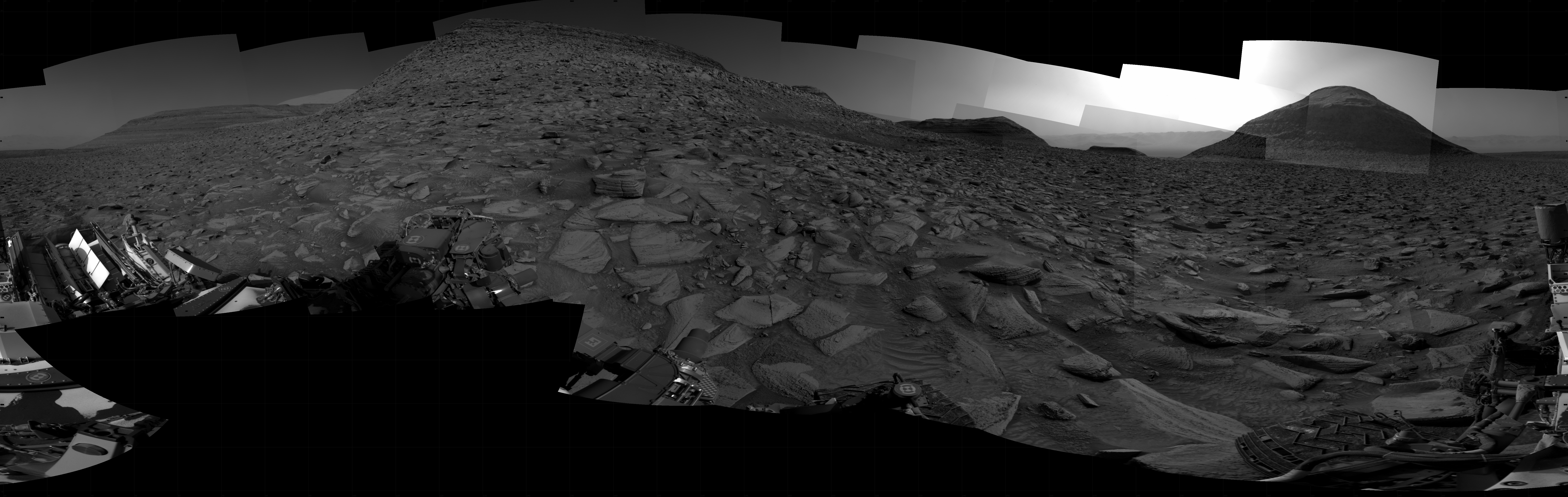 A grayscale panorama of the Martian surface shows a wide field of flat, dark gray terrain dotted with flat, angular, medium-sized rocks stretching into the distance where several hilly features rise from the ground, left to right on the horizon. The largest is a trapezoid-shaped mesa at center left, while the next in size is a smaller, pyramid-shaped hill toward the right side of the frame, silhouetted by a bright light in the sky behind it. Four other features appear smaller and more distant. Portions of the Curiosity rover are visible at the bottom of the image, including one wheel visible in the bottom center of the frame and two others in the lower right corner.