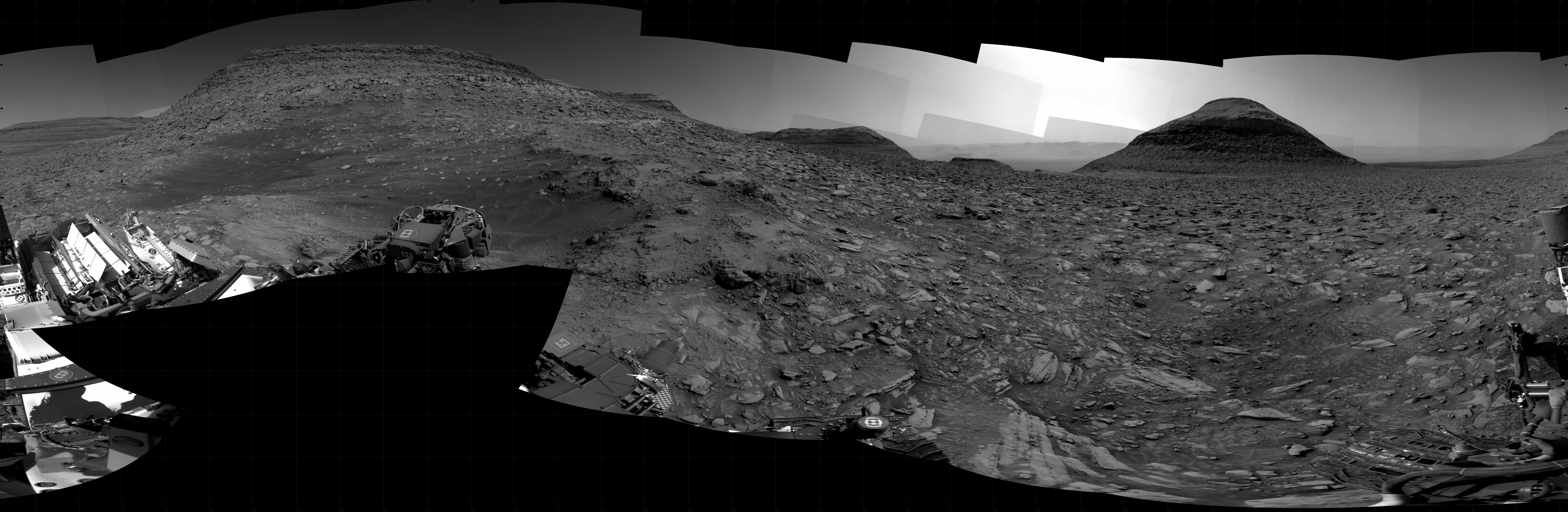 Alt text: A grayscale panorama of the Martian surface, possibly at night or in twilight, shows a wide field of flat, dark gray terrain dotted with flat, angular, medium-sized rocks, lighter gray, stretching into the distance where a pyramid-shaped hill rises from the ground on the horizon, just to the right of image center. A bright light behind the hill illuminates a small part of the sky. Another outcropping, a larger mesa, rises from the ground near the left edge of the frame. Portions of the Curiosity rover are visible at the bottom of the image, including a wheel visible in the lower left of the frame, and another at bottom right.
