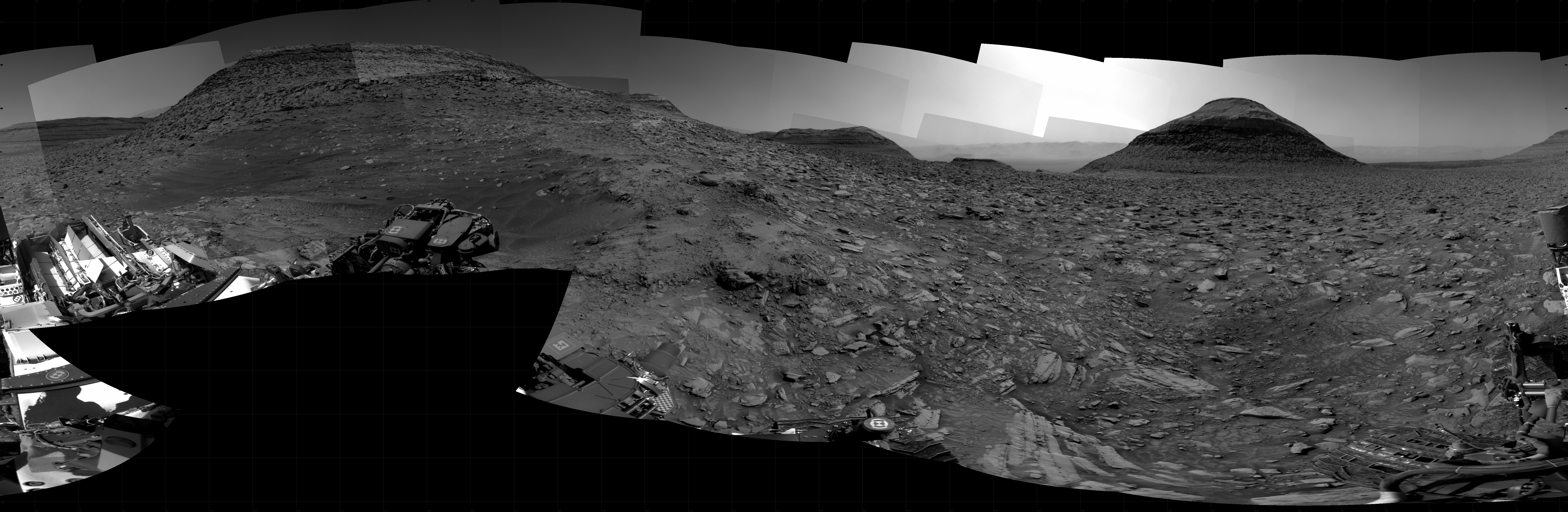 A grayscale panorama of the Martian surface, possibly at night or in twilight, shows a wide field of flat, dark gray terrain dotted with flat, angular, medium-sized rocks, lighter gray, stretching into the distance where a pyramid-shaped hill rises from the ground on the horizon, just to the right of image center. A bright light behind the hill illuminates a small part of the sky. To the left, a larger butte rises from the ground, looking like a pyramid with the top half sheared off. Portions of the Curiosity rover are visible along the bottom of the image, including wheels in the bottom center of the frame and lower-right corner.