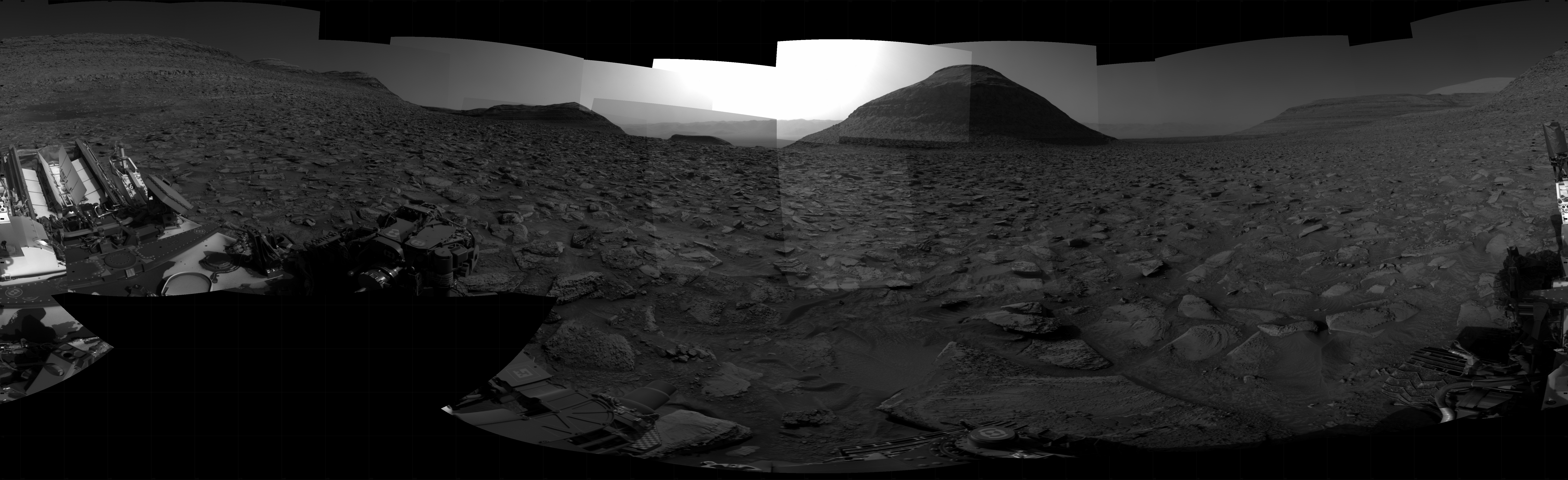 A grayscale panorama of the Martian surface, possibly at night or in twilight, shows a wide field of flat, dark gray terrain dotted with flat, angular, medium-sized rocks, lighter gray, stretching into the distance where a pyramid-shaped hill rises from the ground on the horizon, just to the right of image center. A bright light behind the hill illuminates a small part of the sky. Portions of the Curiosity rover are visible at the bottom of the image, including a wheel visible in the bottom center of the frame, and another in the lower right.