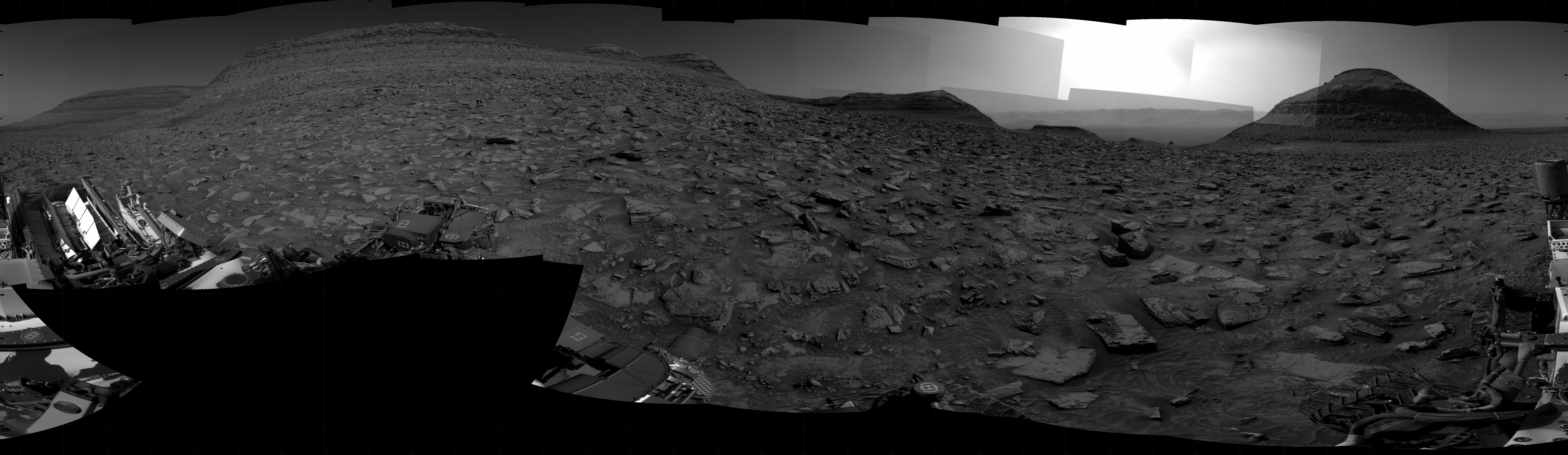 A grayscale panorama of the Martian surface, possibly at night or in twilight, shows a wide field of flat, dark gray terrain dotted with flat, angular, medium-sized rocks, lighter gray, stretching into the distance where a pyramid-shaped hill rises from the ground on the horizon, just to the right of image center. A bright light behind the hill illuminates a small part of the sky. Portions of the Curiosity rover are visible at the bottom of the image, including a wheel visible in the bottom center of the frame, and another two in the lower right.
