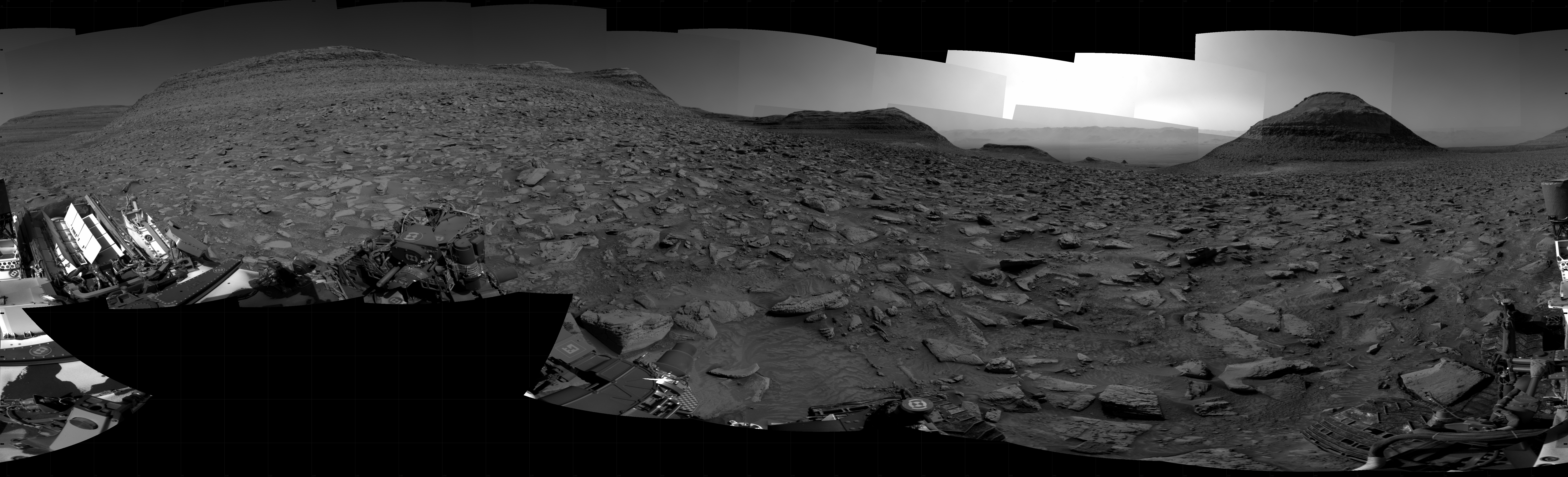 A grayscale panorama of the Martian surface, possibly at night or in twilight, shows a wide field of flat, dark gray terrain dotted with flat, angular, medium-sized rocks, lighter gray, stretching into the distance where a pyramid-shaped hill rises from the ground on the horizon, just to the right of image center. A bright light behind the hill illuminates a small part of the sky. On the far left is a large butte that resembles a pyramid with the top half sliced off. In between are three smaller hills on the horizon. Portions of the Curiosity rover are visible at the bottom of the image, including a wheel visible in the bottom center of the frame, and another two in the lower right.