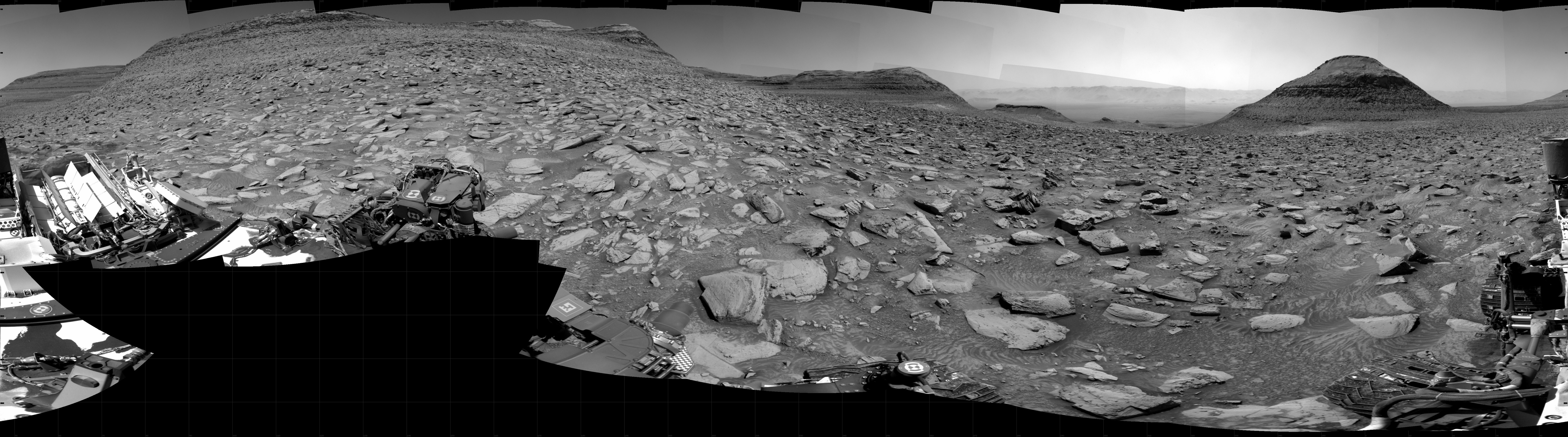 A grayscale panorama of the Martian surface, brightly lit, shows a wide field of flat, dark gray terrain dotted with flat, angular, medium-sized rocks, lighter gray, stretching into the distance where a pyramid-shaped hill rises from the ground on the horizon, just to the right of image center. To its left. smaller hills at varying distances rise up, and at far left a larger butte rises from the ground, looking like a pyramid with the top half sheared off. Portions of the Curiosity rover are visible along the bottom of the image, including a wheel in the bottom center of the frame and two others in the lower-right corner.