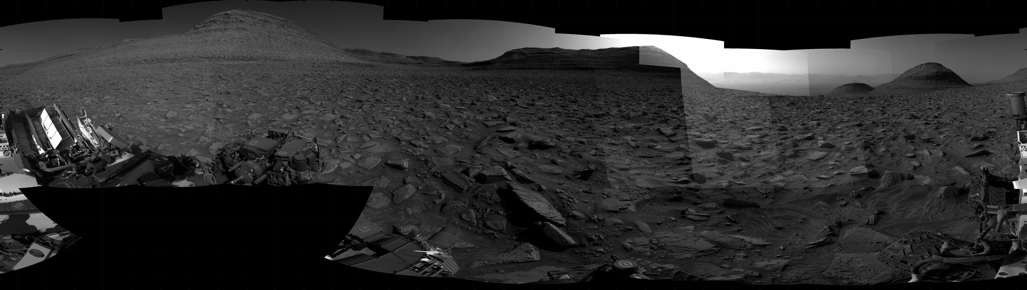 A grayscale panorama of the Martian surface, possibly at night or in twilight, shows a wide field of flat, dark gray terrain dotted with flat, angular, medium-sized rocks stretching into the distance, where a pair of gently sloped, flat-topped hills rise from the ground on the horizon. Behind the one at the right, a bright light behind it illuminates part of the sky, while the rest of the scene is fairly dark. Portions of the Curiosity rover are visible along the bottom of the image, including a wheel at the bottom center of the frame, and two others in the lower-right corner.