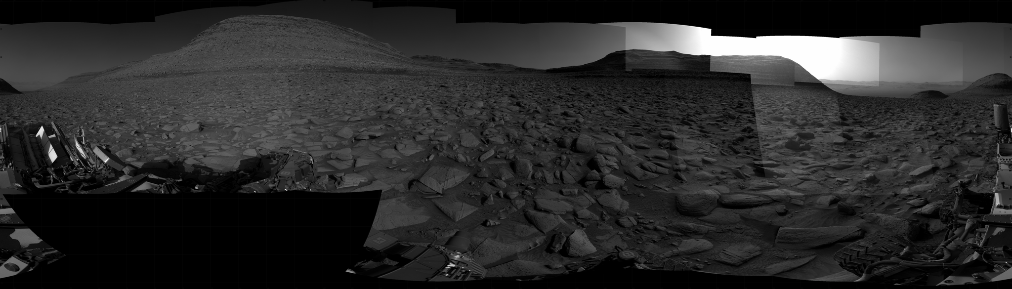 A grayscale panorama of the Martian surface, possibly at night or in twilight, shows a wide field of flat, dark gray terrain dotted with flat, angular, medium-sized rocks stretching into the distance, where a pair of gently sloped, flat-topped buttes rise from the ground on the horizon. Behind the one at the right, a bright light behind it illuminates part of the sky, while the rest of the scene is fairly dark. A smaller, pyramid-shaped hill is at the right edge of the image. Portions of the Curiosity rover are visible along the bottom of the image, including a wheel at the bottom center of the frame, and two others in the lower-right corner.
