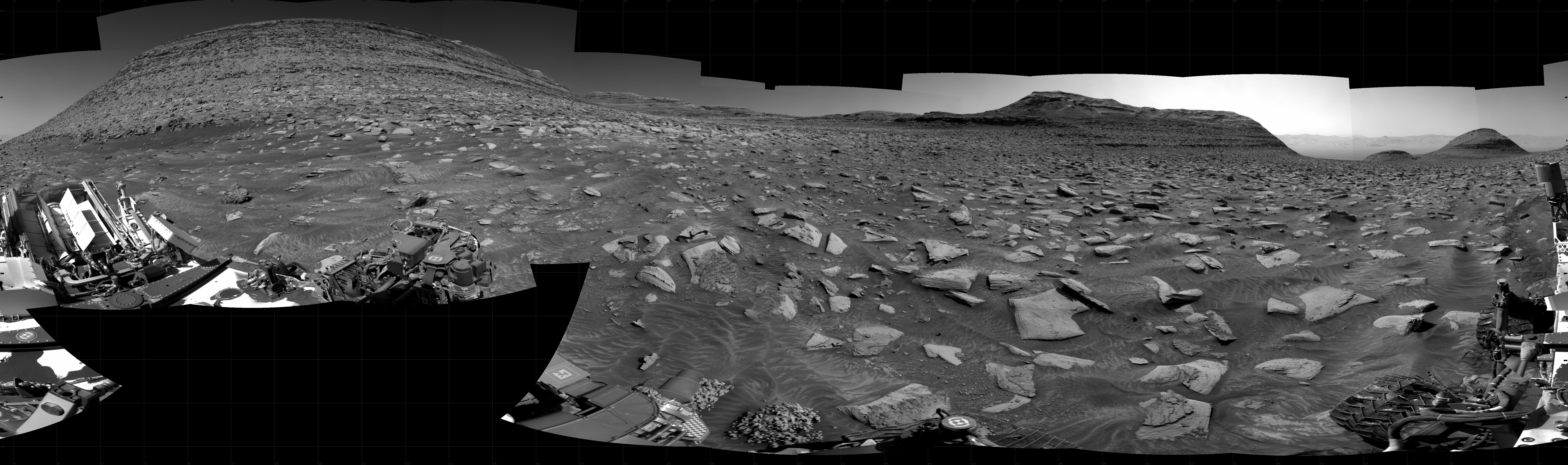 A grayscale panorama of the Martian surface shows a wide field of flat, dark gray terrain dotted with flat, angular, medium-sized rocks stretching into the distance where several features rise from the ground. At left a very large mesa, nearly reaching the top of the frame. In the middle, a smaller mesa, a little farther away. And at the far right, two much smaller, gently sloped hills, looking to be even farther from the rover. Portions of the Curiosity rover are visible at the bottom of the image, including one wheel visible in the bottom center of the frame and two others in the lower right corner.