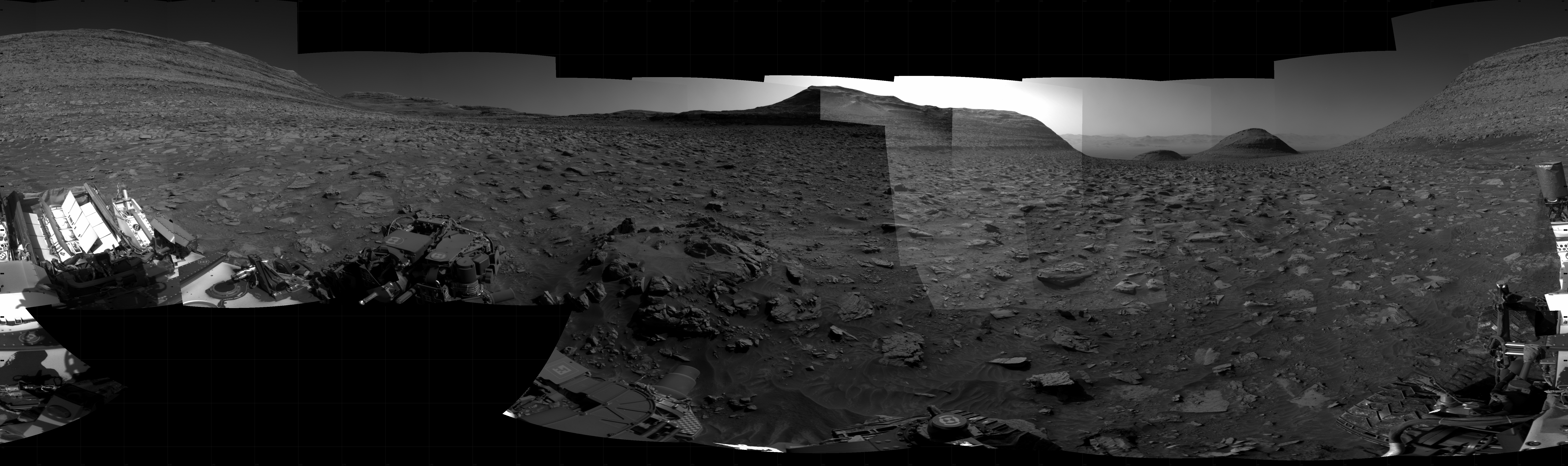 A grayscale panorama of the Martian surface, possibly at night or in twilight, shows a wide field of flat, dark gray terrain dotted with flat, angular, medium-sized rocks stretching into the distance where several features rise from the ground. At left is a very large mesa, nearly reaching the top of the frame. In the middle, a smaller mesa, a little farther away — a very bright light source illuminates the sky above this. To the right of that, two much smaller, gently sloped hills, looking to be even farther from the rover. And at the far right, the other half of the largest mesa, at far left, rises up to complete the 360-degree view. Portions of the Curiosity rover are visible at the bottom of the image, including one wheel visible in the bottom center of the frame and two others in the lower right corner.