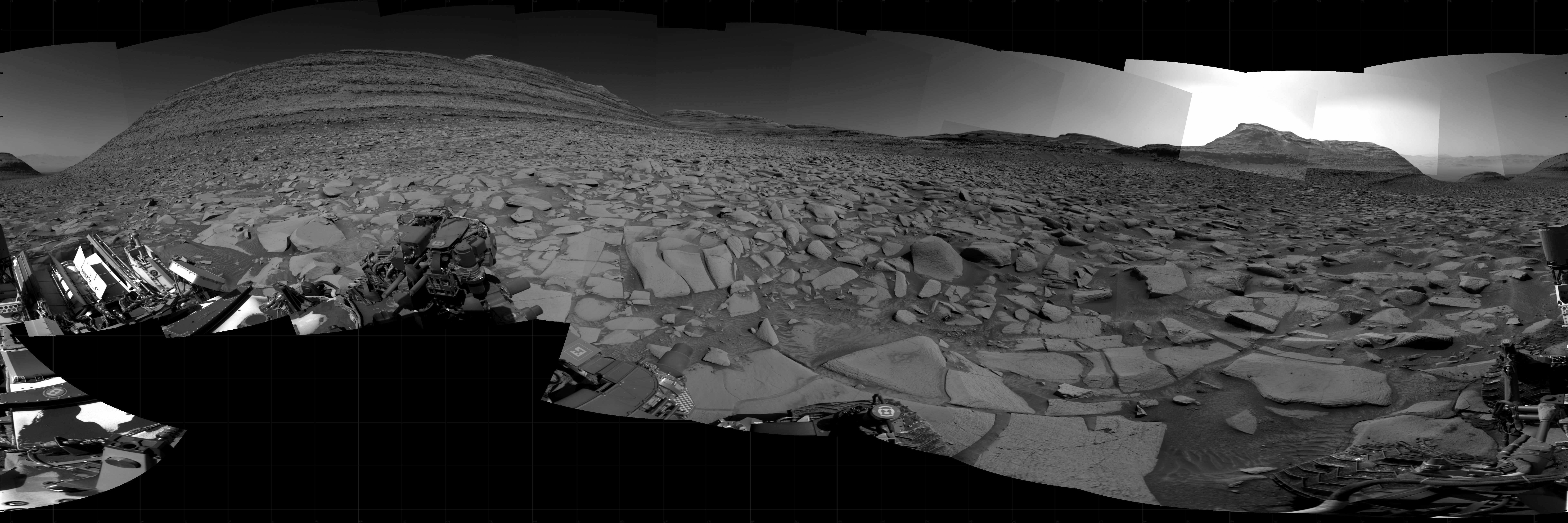 A grayscale panorama of the Martian surface, possibly at night or in twilight, shows a wide field of flat, dark gray terrain dotted with flat, angular, medium-sized rocks stretching into the distance where a couple of features rise from the ground. At left is a very large mesa, nearly reaching the top of the frame. At right, smaller hills lead up to a medium-sized mesa, behind which a very bright light source illuminates the sky above. Portions of the Curiosity rover are visible at the bottom of the image, including one wheel visible in the bottom center of the frame and two others in the lower right corner.
