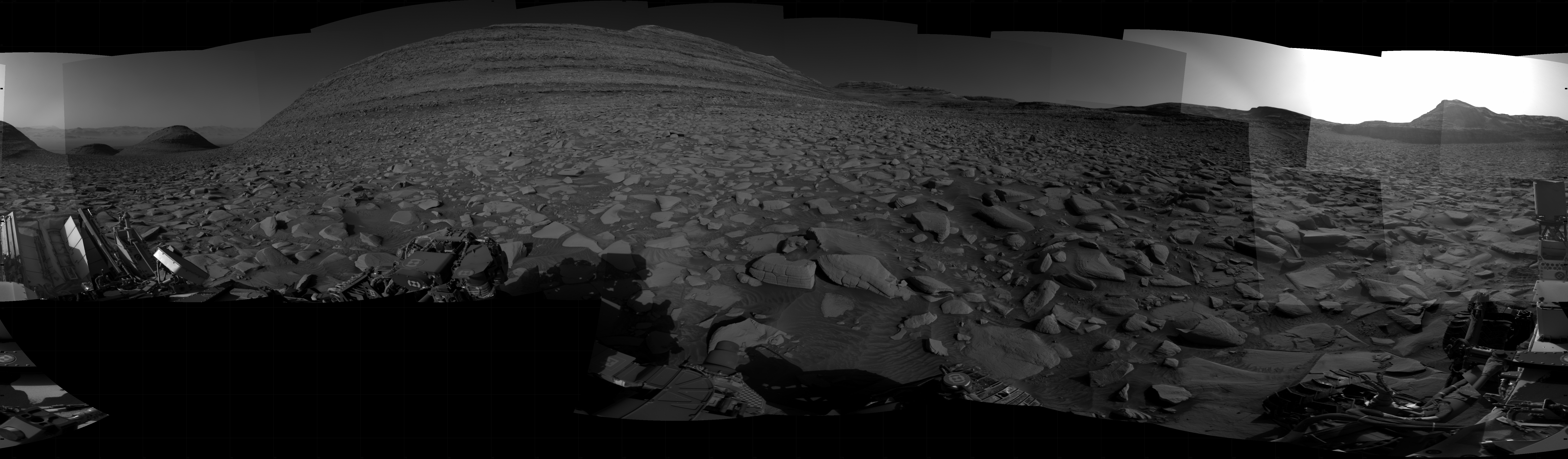 A grayscale panorama of the Martian surface, possibly at night or in twilight, shows a wide field of flat, dark gray terrain dotted with flat, angular, medium-sized rocks stretching into the distance where features rise from the ground. At center-left is a very large mesa, nearly reaching the top of the frame. At far right, a small, pyramid shaped hill is backlit by a very bright light source that illuminates the sky above. Portions of the Curiosity rover are visible at the bottom of the image, including one wheel visible in the bottom center of the frame and two others in the lower right corner.
