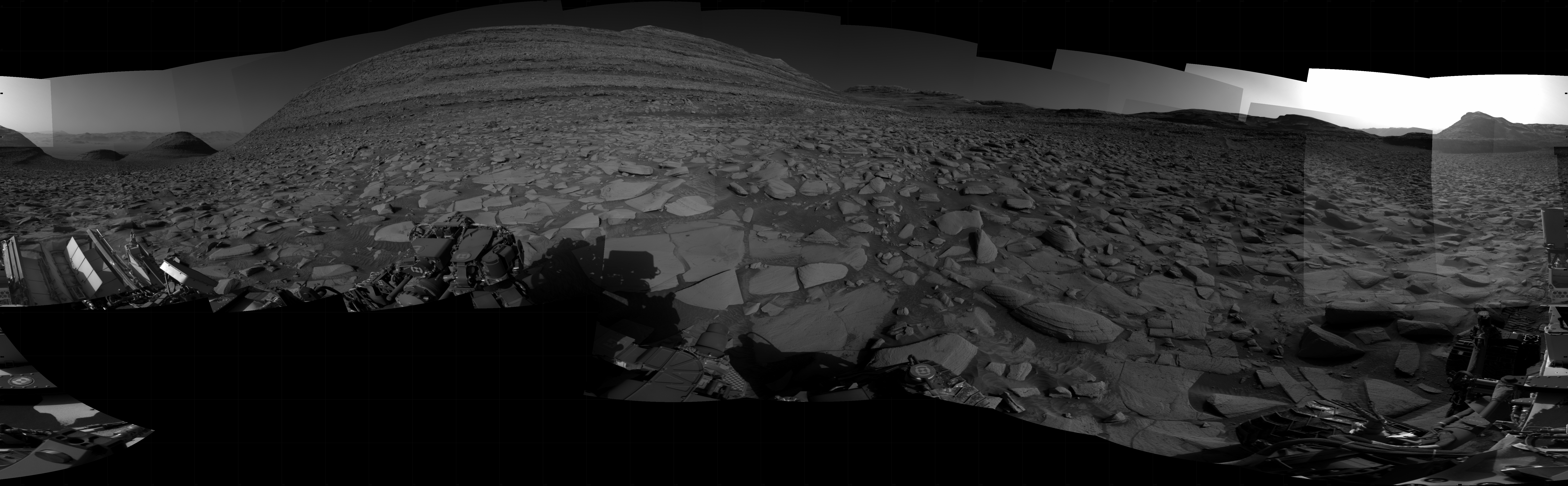 A grayscale panorama of the Martian surface, possibly at night or in twilight, shows a wide field of flat, dark gray terrain dotted with flat, angular, medium-sized rocks stretching into the distance where features rise from the ground. At center-left is a very large mesa, nearly reaching the top of the frame.Three small pyramid-shaped hills are to the left of that. At far right, a small, pyramid shaped hill is backlit by a very bright light source that illuminates the sky above. Portions of the Curiosity rover are visible at the bottom of the image, including one wheel visible in the bottom center of the frame and two others in the lower right corner.