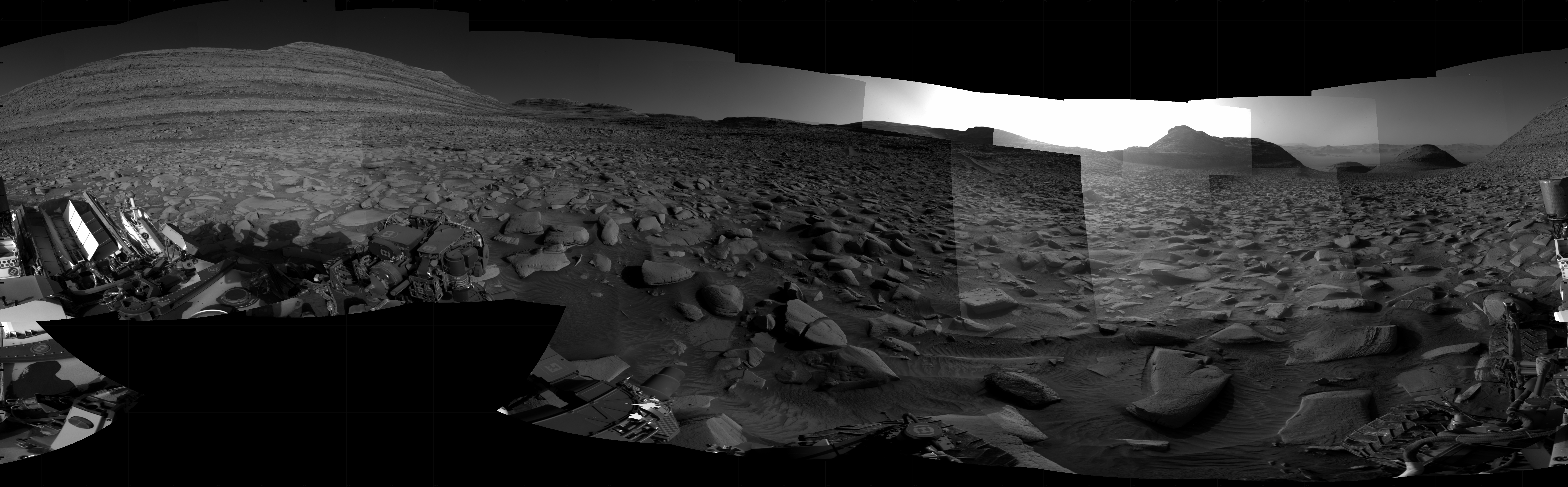 A grayscale panorama of the Martian surface, possibly at night or in twilight, shows a wide field of flat, dark gray terrain dotted with flat, angular, medium-sized rocks stretching into the distance where features rise from the ground. At left is a very large mesa, nearly reaching the top of the frame. At right, several small, pyramid shaped hills are backlit by a very bright light source that illuminates the sky above. Portions of the Curiosity rover are visible at the bottom of the image, including one wheel visible in the bottom center of the frame and two others in the lower right corner.
