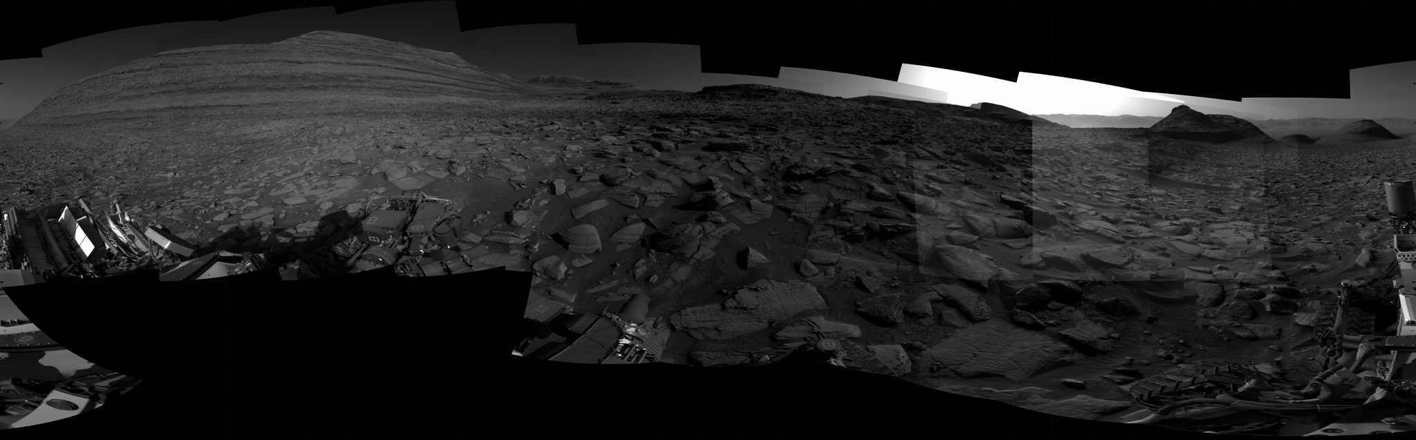 A grayscale panorama of the Martian surface, possibly at night or in twilight, shows a wide field of flat, dark gray terrain dotted with flat, angular, medium-sized rocks stretching into the distance where features rise from the ground. At left is a very large mesa, nearly reaching the top of the frame. At right, several small, pyramid shaped hills are backlit by a very bright light source that illuminates the sky above. Portions of the Curiosity rover are visible at the bottom of the image, including one wheel visible in the bottom center of the frame and two others in the lower right corner.