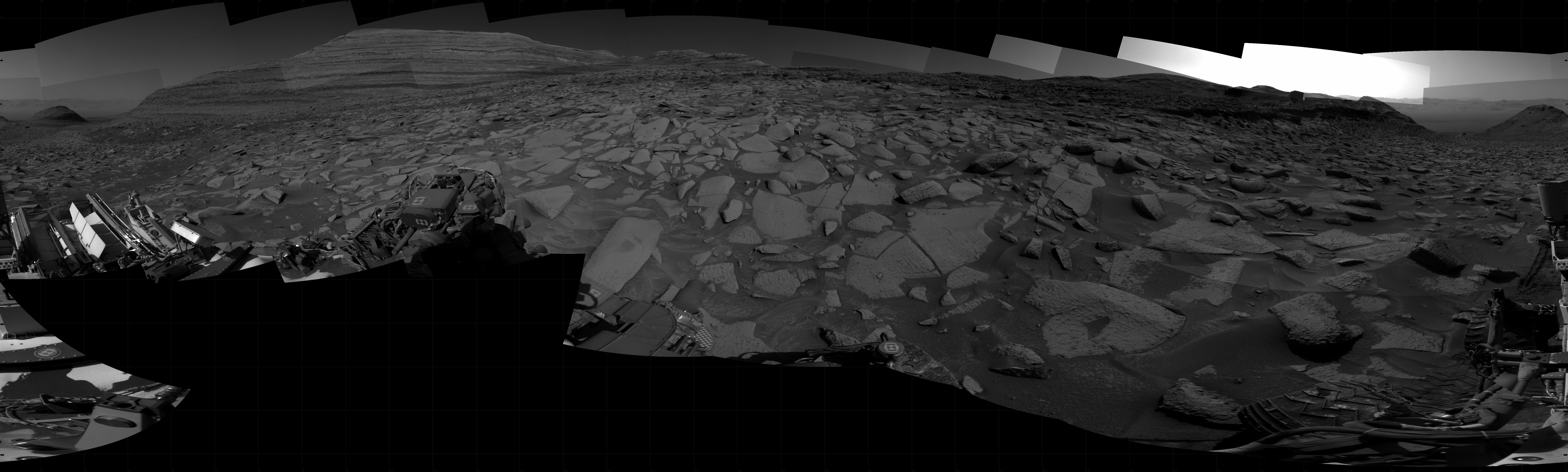 Panoramic black-and-white image of the rocky Martian landscape taken by the Curiosity rover, showing rugged terrain, layered hills in the distance, and parts of the rover's structure in the foreground.