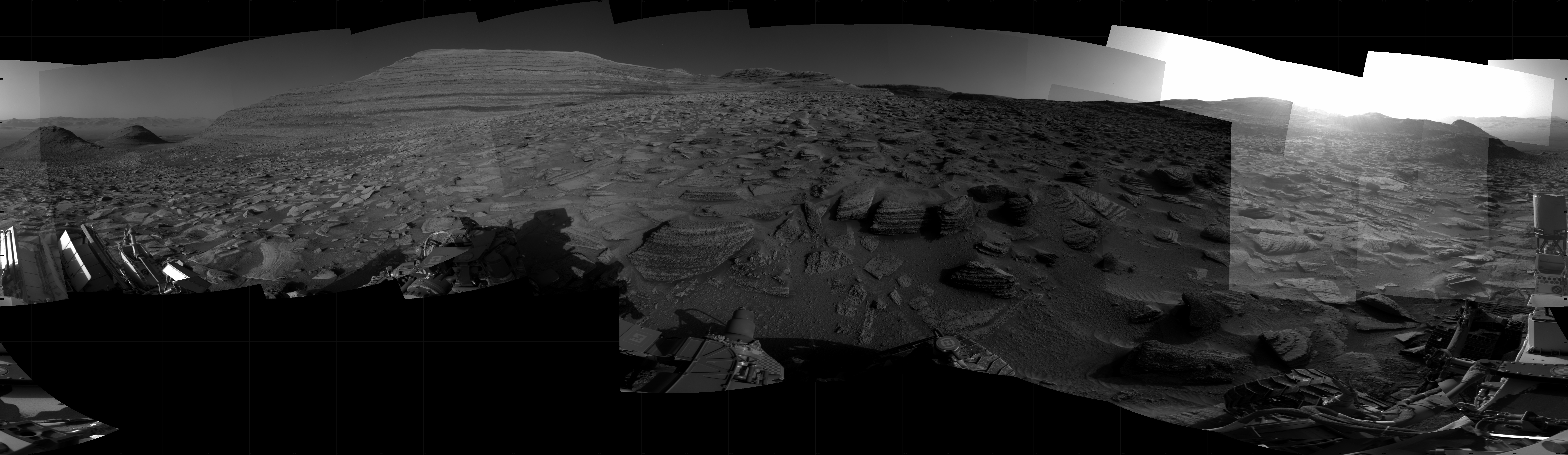 Panoramic black-and-white image of the rocky Martian landscape taken by the Curiosity rover, showing rugged terrain, layered hills in the distance, and parts of the rover's structure in the foreground.