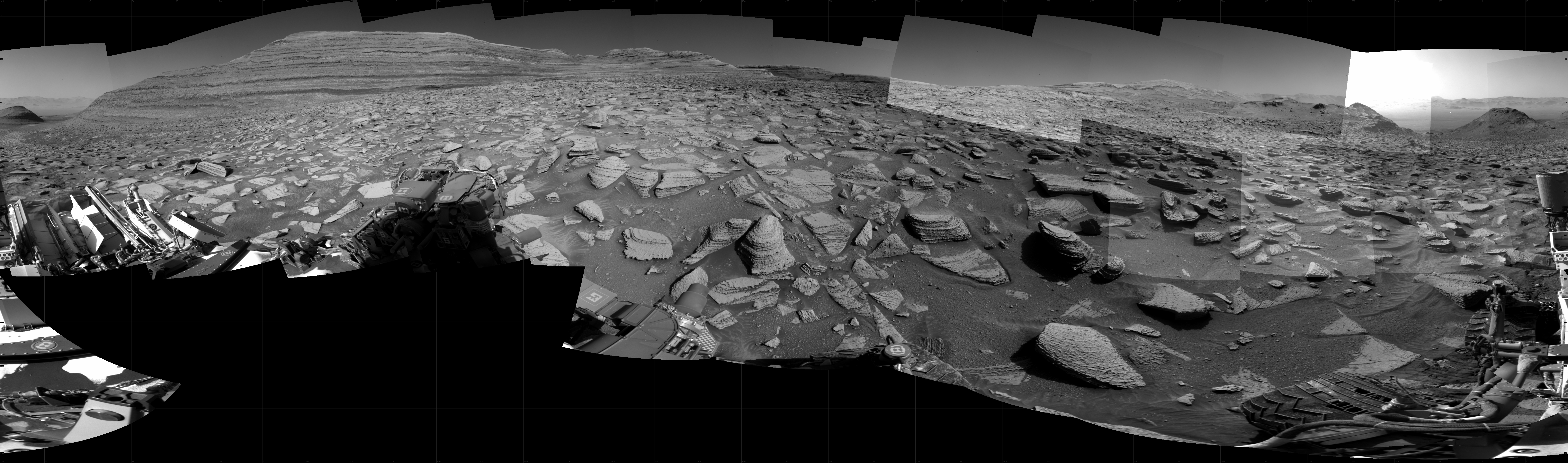 Panoramic black-and-white image of the rocky Martian landscape taken by the Curiosity rover, showing rugged terrain, layered hills in the distance, and parts of the rover's structure in the foreground.